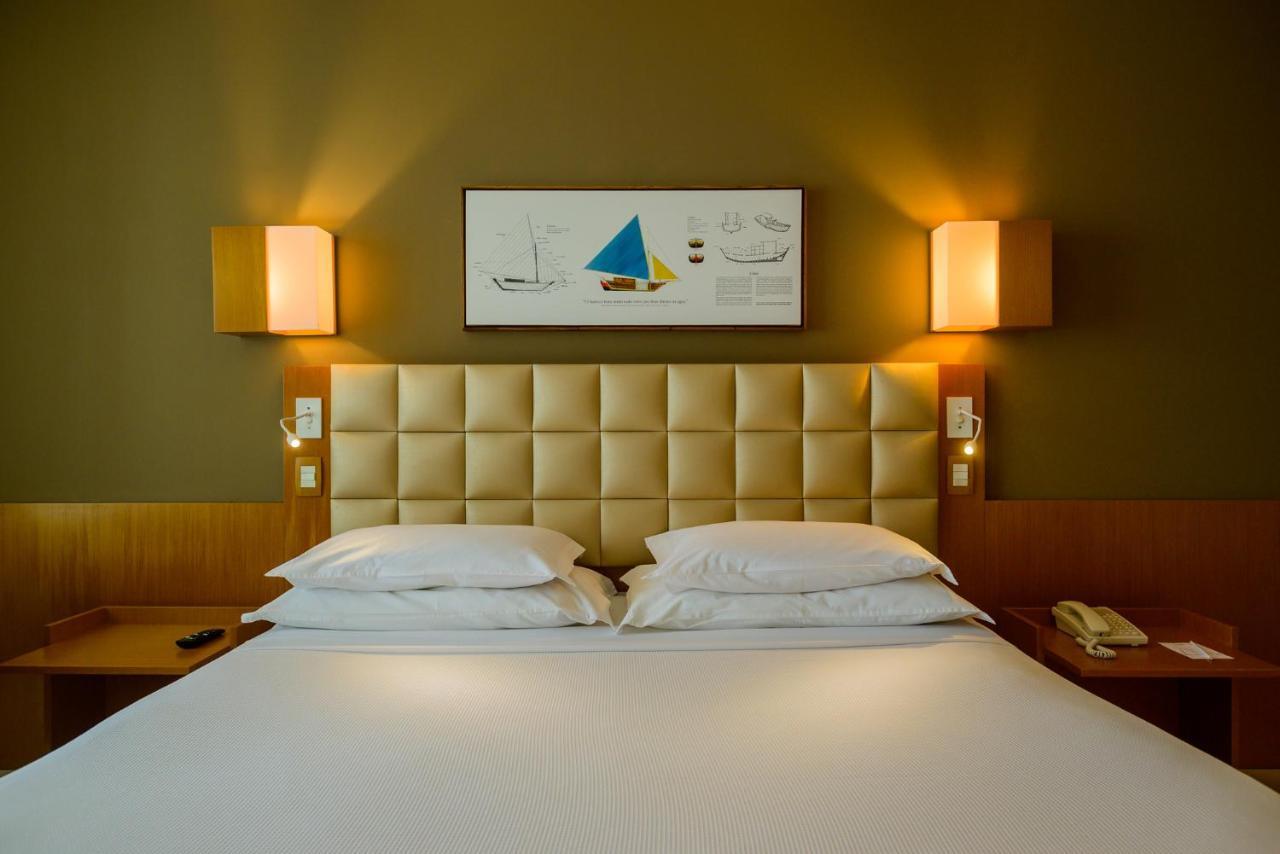 Hotel Luzeiros Sao Luis Экстерьер фото The photo shows a modern hotel room featuring a large bed with a tufted beige headboard. There are two white pillows on the bed and a smooth white bedspread. On the wall above the bed, there's a framed artwork with a colorful sailboat and nautical de