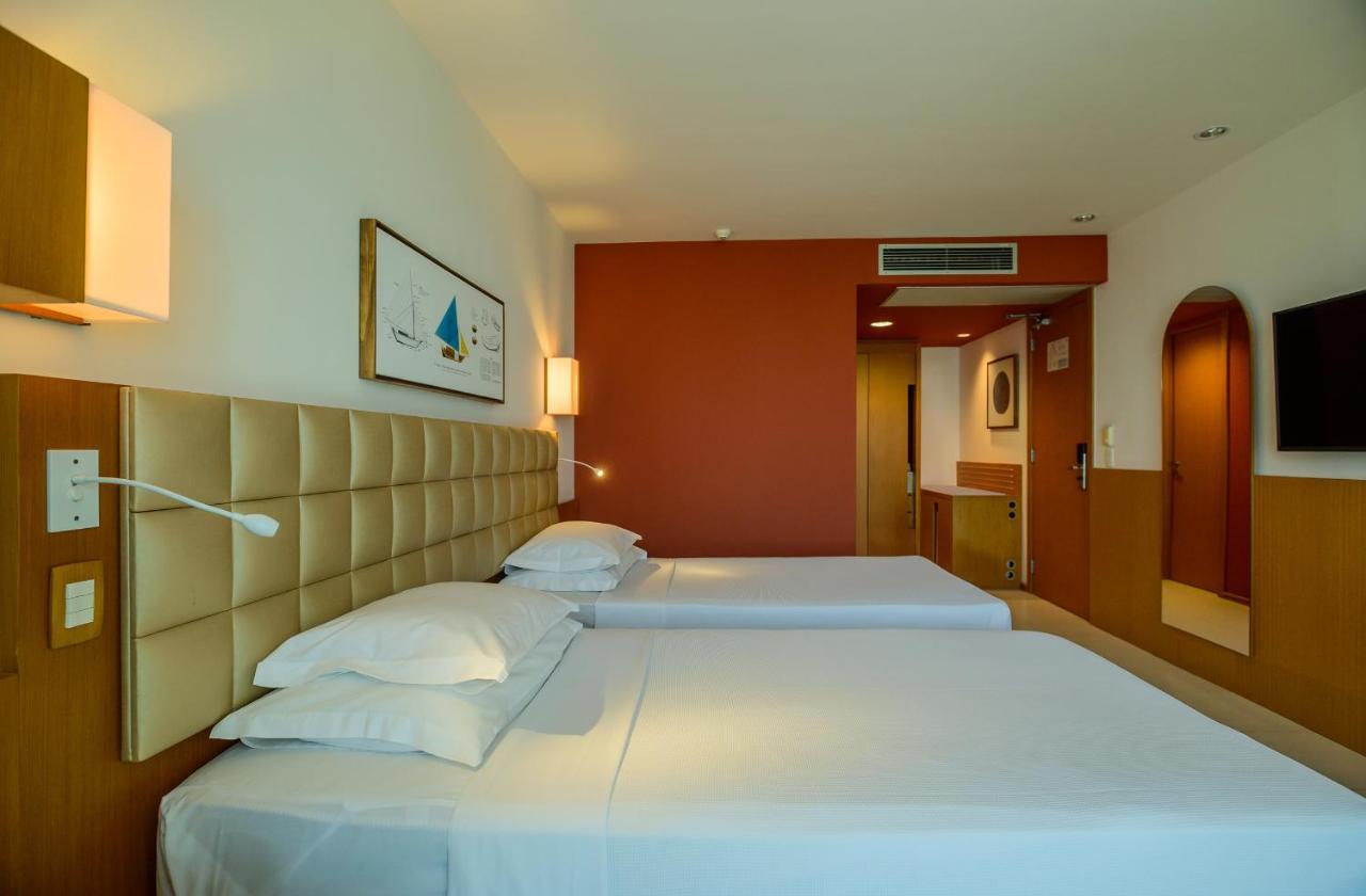 Hotel Luzeiros Sao Luis Экстерьер фото The photo shows a hotel room featuring two beds with white linens and pillows. The beds are positioned next to a beige headboard. There is a wall with a reddish hue that contrasts with the lighter colors of the bedding. A piece of artwork is hanging 
