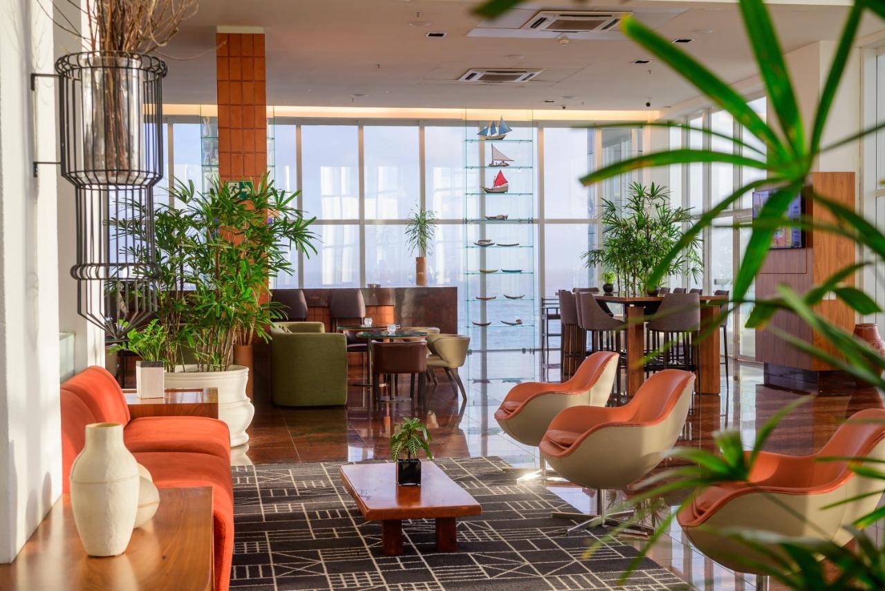 Hotel Luzeiros Sao Luis Экстерьер фото The photo shows a modern, stylish indoor lounge area with a bright and airy atmosphere. There are several seating arrangements, including chairs with orange upholstery and green sofas. A wooden coffee table is centered on a patterned rug. The space f