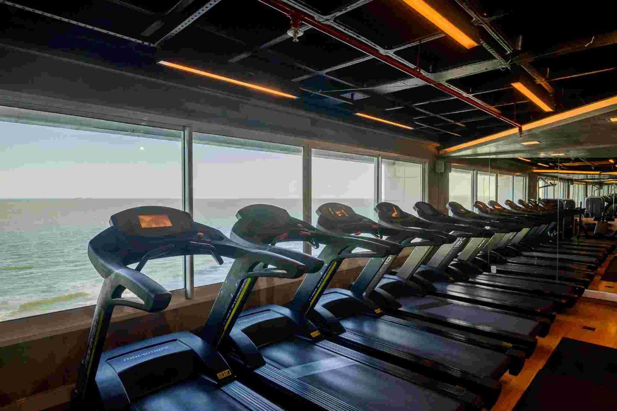 Hotel Luzeiros Sao Luis Экстерьер фото The photo shows a row of treadmills lined up inside a gym or fitness center. The treadmills are adjacent to large windows that provide a view of the ocean or a body of water. The area has modern lighting fixtures and a contemporary design, creating a