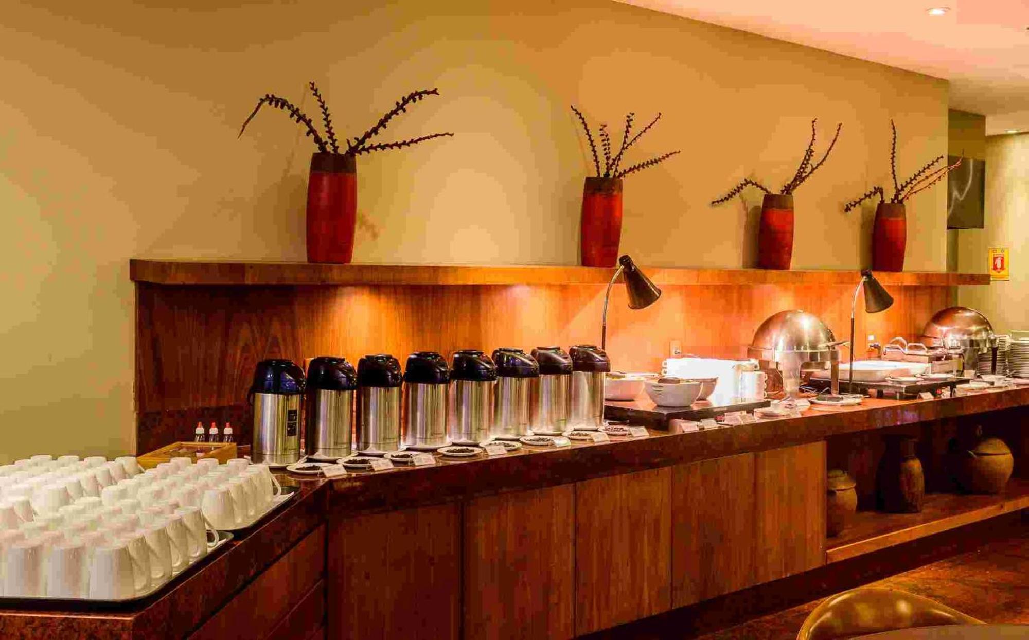 Hotel Luzeiros Sao Luis Экстерьер фото The photo shows a buffet setup in a dining area. There is a long wooden counter that features several items:

1. **Beverages**: Multiple stainless steel coffee or tea containers are lined up, suggesting a beverage station.
2. **Dishware**: There are 