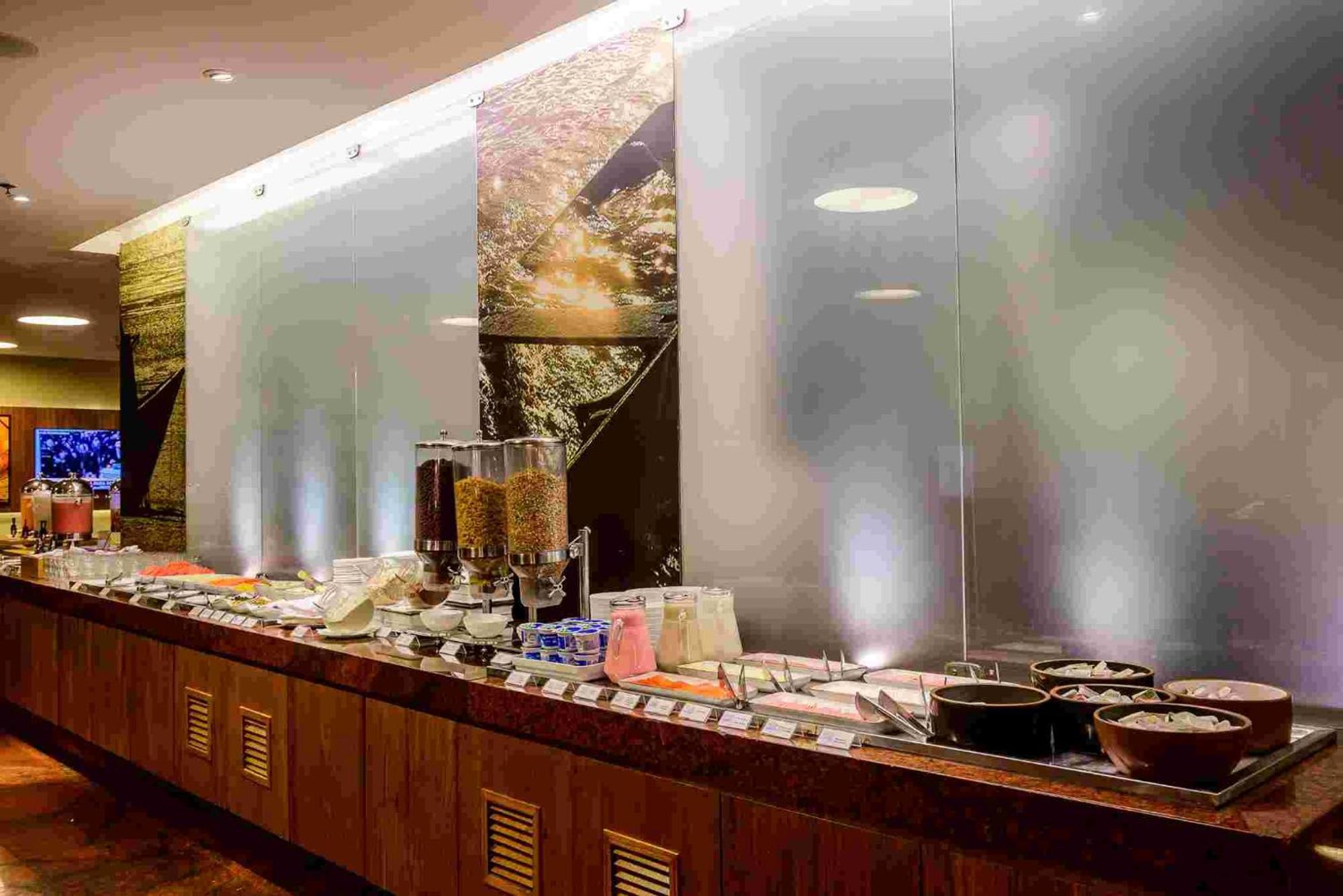Hotel Luzeiros Sao Luis Экстерьер фото The photo shows a buffet setup in a restaurant or dining area. There is a long wooden countertop displaying a variety of food items arranged on it, including containers and serving dishes. Behind the buffet, there are large panels or screens, which m