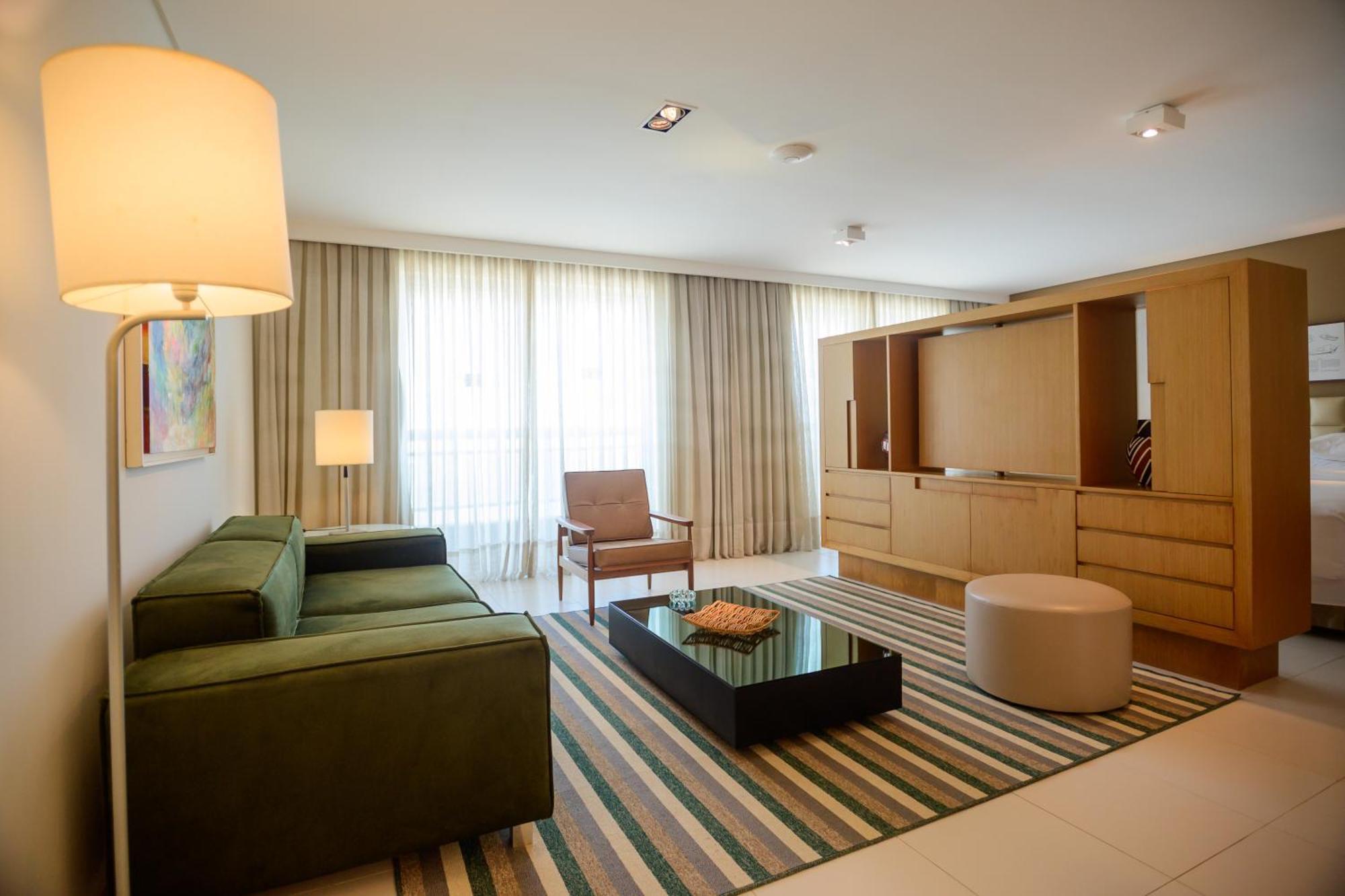 Hotel Luzeiros Sao Luis Экстерьер фото The photo shows a modern living room with a clean and minimalist design. There is a green sectional sofa positioned against one wall, complemented by a brown armchair nearby. A coffee table with a black surface sits in the center of the room, with a 