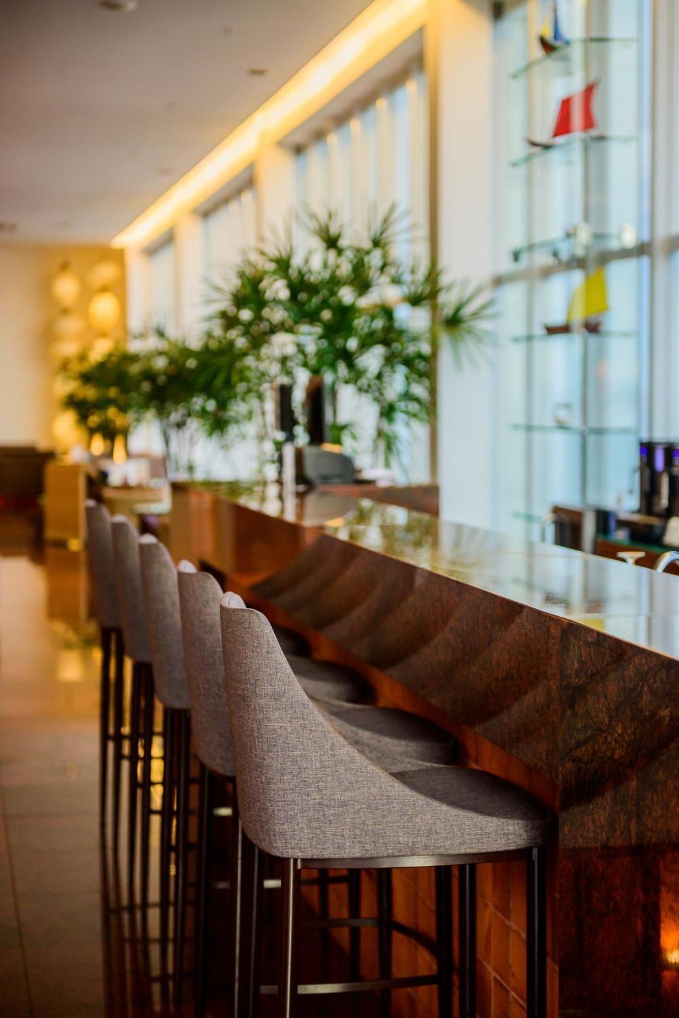 Hotel Luzeiros Sao Luis Экстерьер фото The photo shows a modern bar area with a sleek, polished countertop made of what appears to be stone or marble. There are several bar stools with rounded backs arranged around the counter. In the background, there are lush green plants providing a na