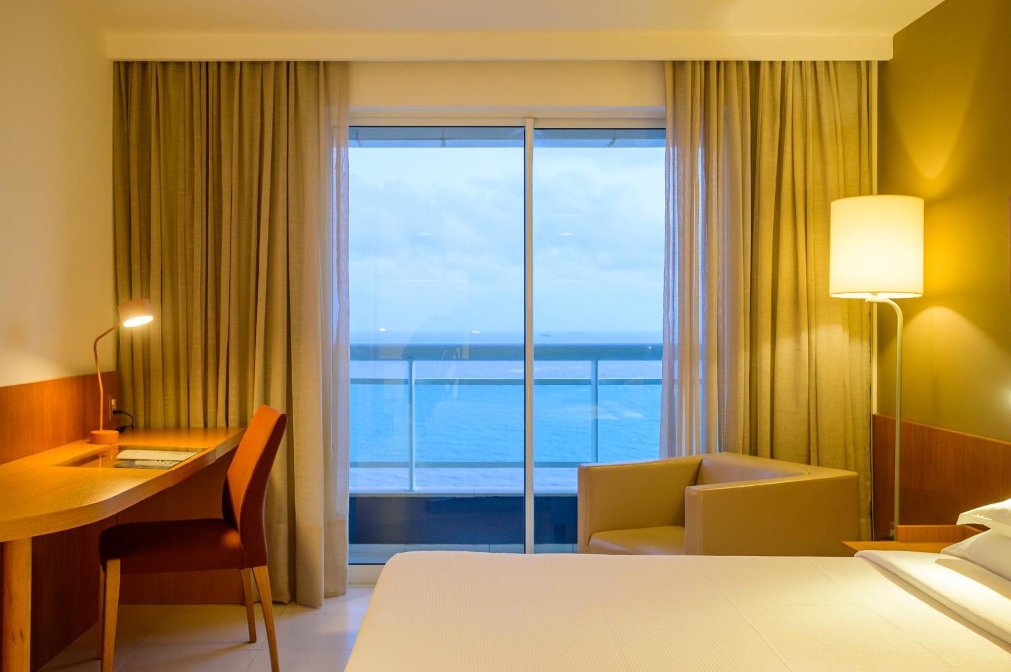 Hotel Luzeiros Sao Luis Экстерьер фото The photo shows a modern hotel room with a minimalist design. There's a large bed with white linens, and beside it, a small desk with a chair, illuminated by a lamp. A cozy armchair is positioned near a large window, which offers a view of the ocean 
