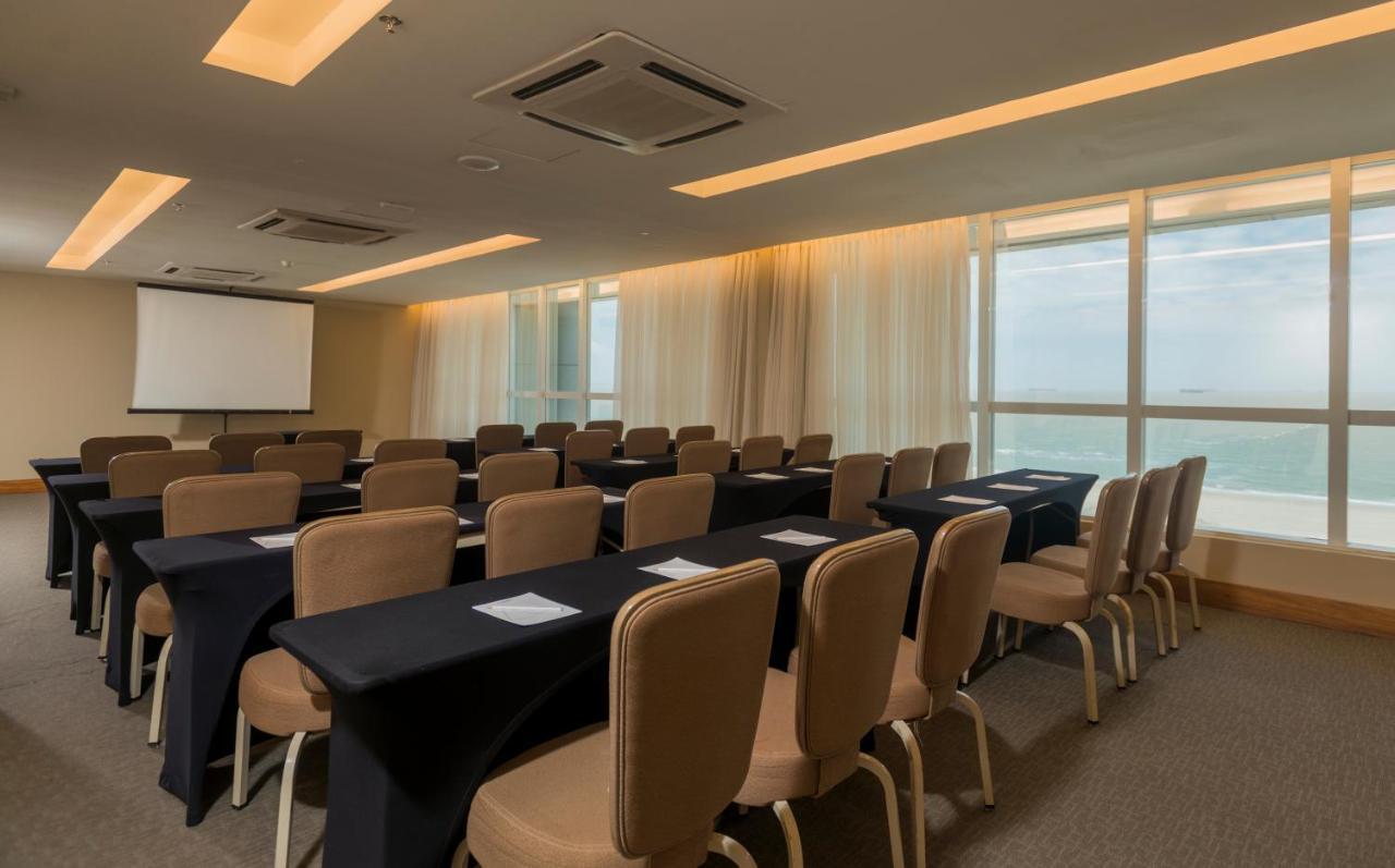 Hotel Luzeiros Sao Luis Экстерьер фото The photo shows a meeting or conference room. There are rows of rectangular tables covered with black tablecloths, each accompanied by beige chairs. A screen for presentations is visible at the front of the room, and large windows let in natural ligh